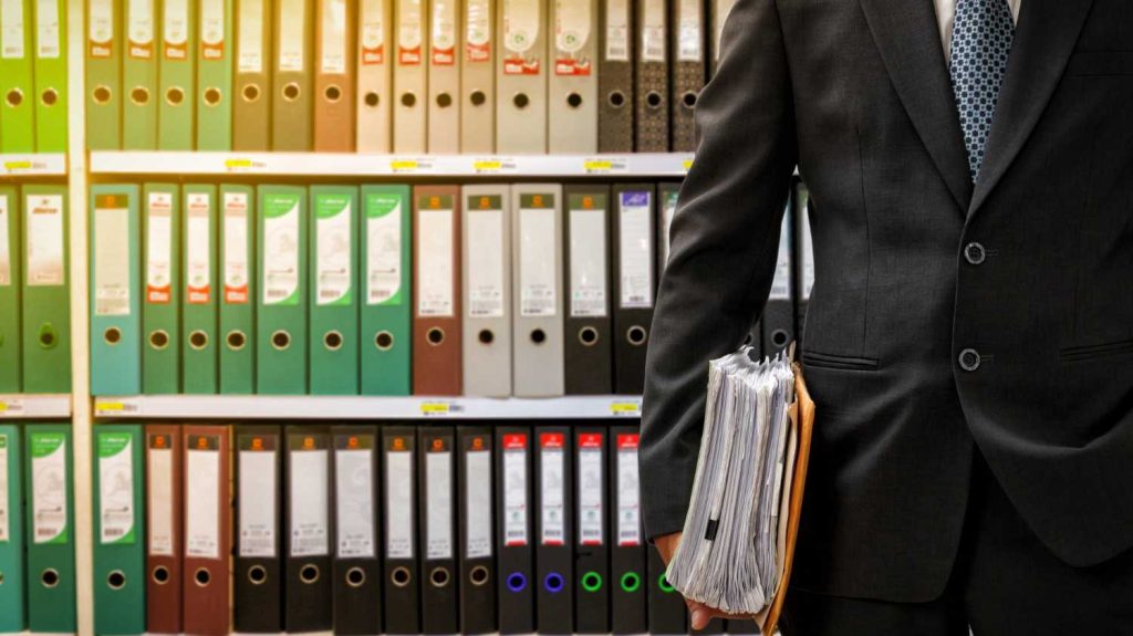 Mężczyzna w garniturze trzyma papierowe dokumenty i segregatory, co nawiązuje do normy DIN 66399 i niszczenia poufnej dokumentacji.