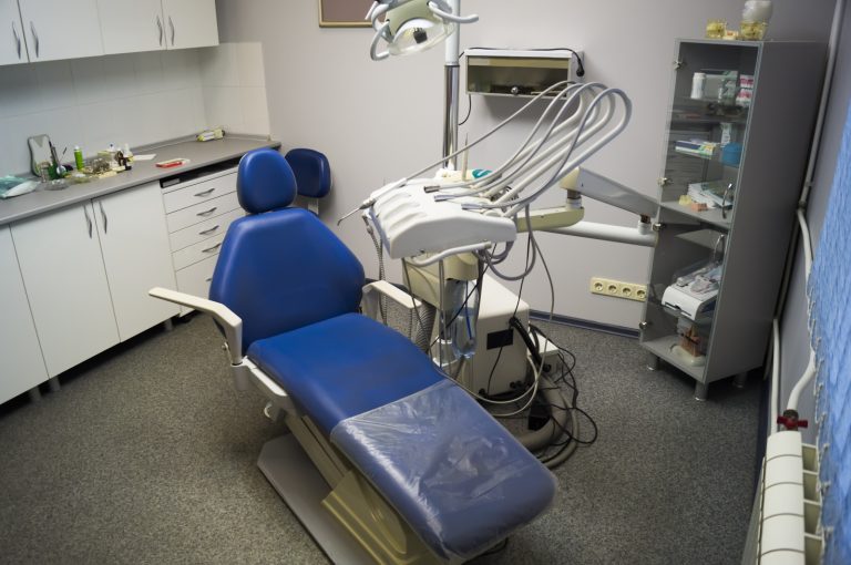 Niebieski fotel stomatologiczny z osprzętem w białej zabudowie i metalowych elementach, widoczne przewody oraz stanowisko pracy w gabinecie.